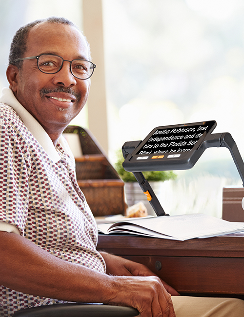 Image of a man seated at table with explorē 8 electronic magnifier on stand. The magnifier displays the text of a magazine underneath the stand.