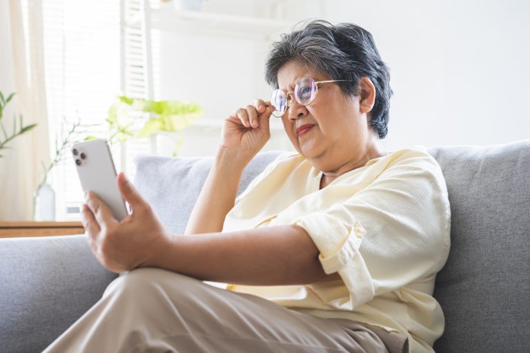 Mature, aged asian woman wearing glasses having vision problem trying to stare text on smart mobile phone screen, eye disease of elderly. Poor eyesight farsightedness concept.