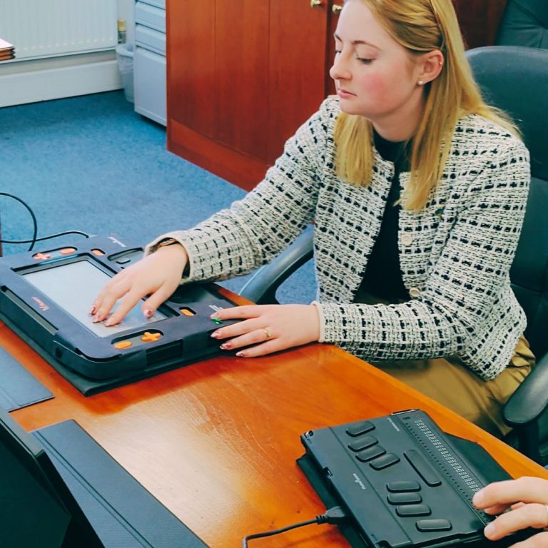 Gen Kitchen MP touching the multi-line braille display of a Monarch. In the foreground is a BrailleNote Touch Plus. In the background a VictorReader Stratus and an Index Basic-D is sitting on a cabinet.