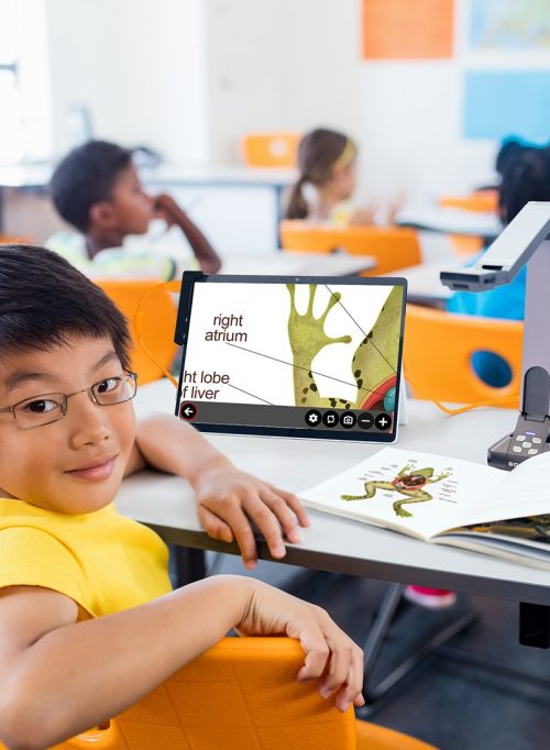 Image of a classroom with a student using a digital tablet connected to the camera to enlarge a book on the biology of a frog.