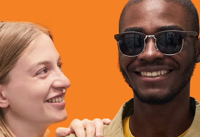 A smiling young woman touching the shoulder of her visually impaired friend wearing sunglasses.